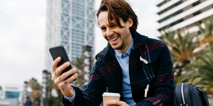 Mann sitzt auf Parkbank, trinkt Kaffee und schaut auf Smartphone.