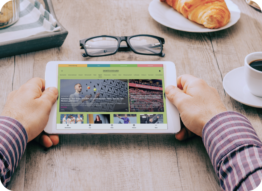 Männerhände, die am Frühstückstisch ein Tablet halten, auf dem die DER STANDARD App zu sehen ist.